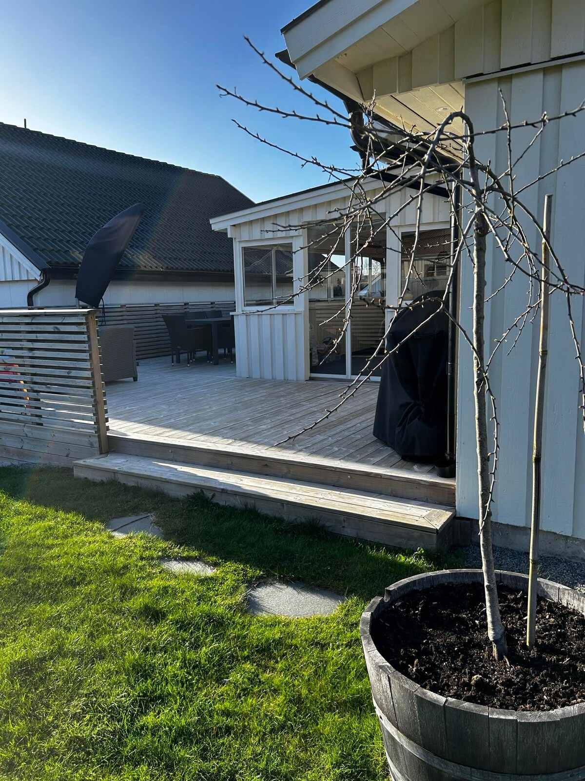 A wooden deck extends from the house, featuring a dining area with a table and chairs. A small tree in a planter adds a touch of greenery, while the sky is clear and the sun casts shadows on the grass. The surrounding area is well-maintained.