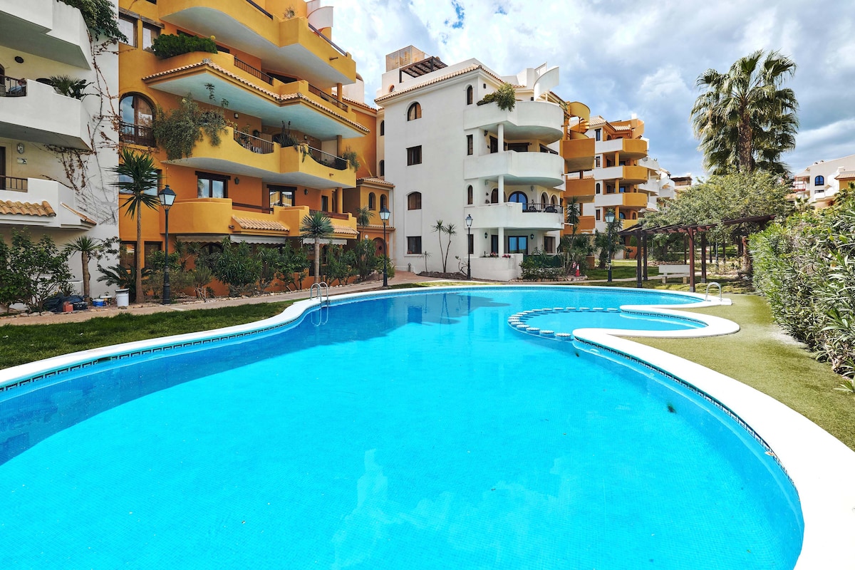 A large swimming pool with clear blue water is surrounded by lush greenery and colorful buildings. A well-maintained garden area features palm trees and seating options, creating an inviting space for relaxation. The layout offers both sunlit and shaded areas for guests.