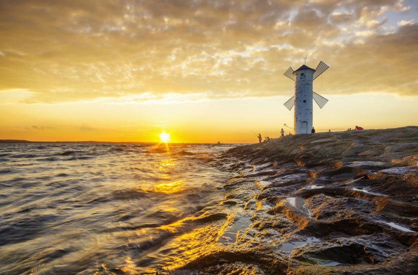 A scenic view of a windmill against a sunset sky, with warm tones reflecting on the water's surface. The shoreline features smooth rocks and gentle waves, creating a serene landscape. Silhouetted figures can be seen enjoying the area, lending a sense of peaceful activity.