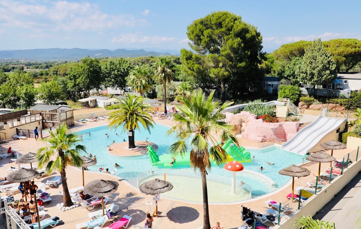 A large outdoor pool area is surrounded by palm trees and lush greenery. Multiple sun loungers line the poolside, and a water slide is visible, adding to the recreational appeal. The scene captures a vibrant atmosphere enjoyed by guests on sunny day.