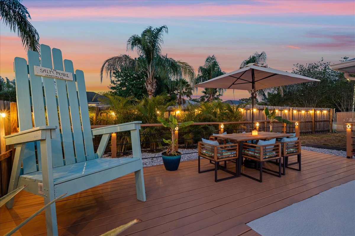 A large blue chair with 'Salty Pirate' signage is positioned on a wooden deck, surrounded by lush greenery and palm trees. A dining area with a table and chairs is shaded by an umbrella, while soft lighting creates an inviting ambiance as the sun sets in the background.
