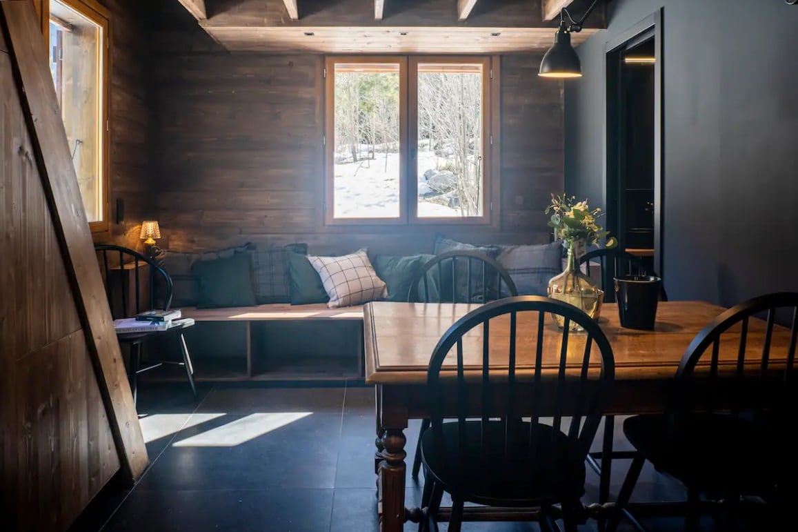 A warm, inviting space features a wooden interior with two large windows allowing natural light to fill the room. A cozy seating area with cushions is adjacent to a dining table, where dark chairs surround a wooden tabletop. Fresh flowers add a touch of nature.
