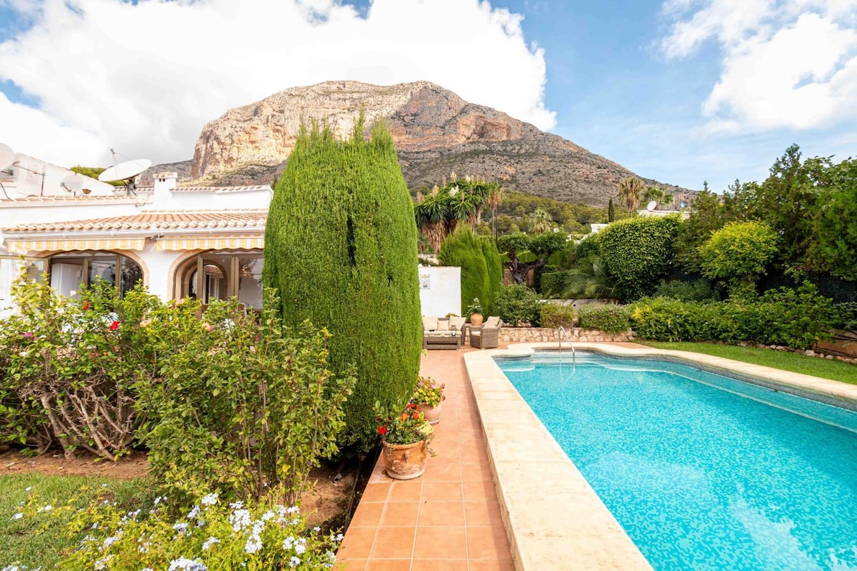 A serene private heated swimming pool is framed by lush greenery and vibrant flowers. The Montgo mountain looms in the background, providing a stunning natural backdrop. Comfortable seating areas are arranged near the pool, inviting relaxation in a tranquil outdoor setting.