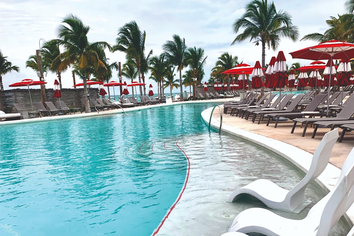 An expansive infinity pool is surrounded by lounge chairs and vibrant red umbrellas. Lush palm trees line the area, providing a tropical ambiance. The clear water reflects the cloudy sky, creating a serene atmosphere for relaxation.