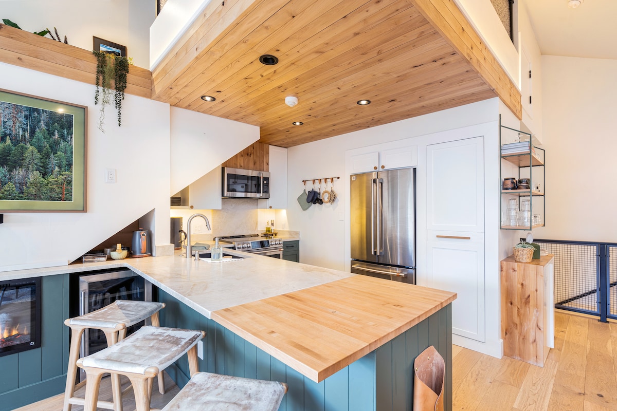The kitchen is designed with modern appliances and a spacious island with a light wood countertop. Soft lighting highlights the wood-paneled ceiling, creating a cozy atmosphere. Bar stools provide seating at the island, while a stylish mix of cabinetry offers ample storage.