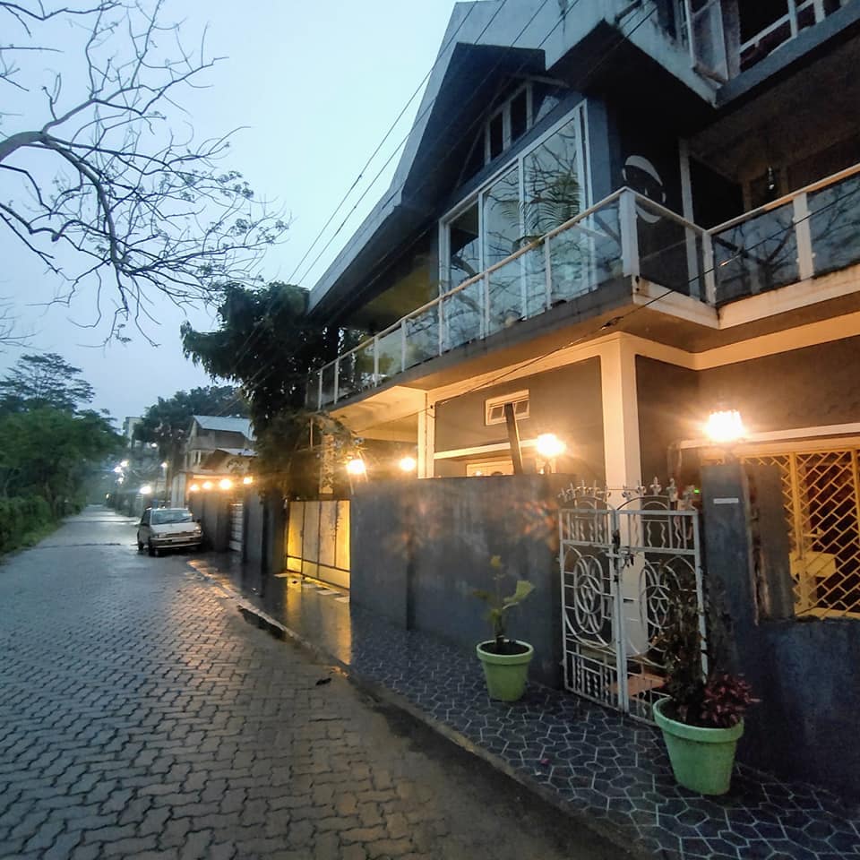A serene street scene is captured at dusk, showcasing a charming building with large windows and a balcony. Soft lights illuminate the pathway lined with potted plants, while a parked car is visible beneath the gentle glow of street lamps.