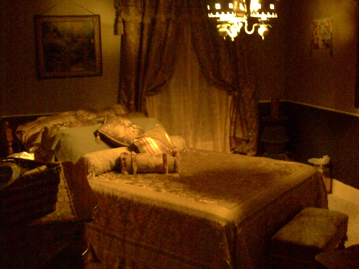 A cozy sleeping area features a spacious bed draped in rich textiles, complemented by decorative pillows. A softly glowing chandelier hangs above, while warm light filters through thick drapes. A painting adorns the wall, contributing to the inviting ambiance of the room.