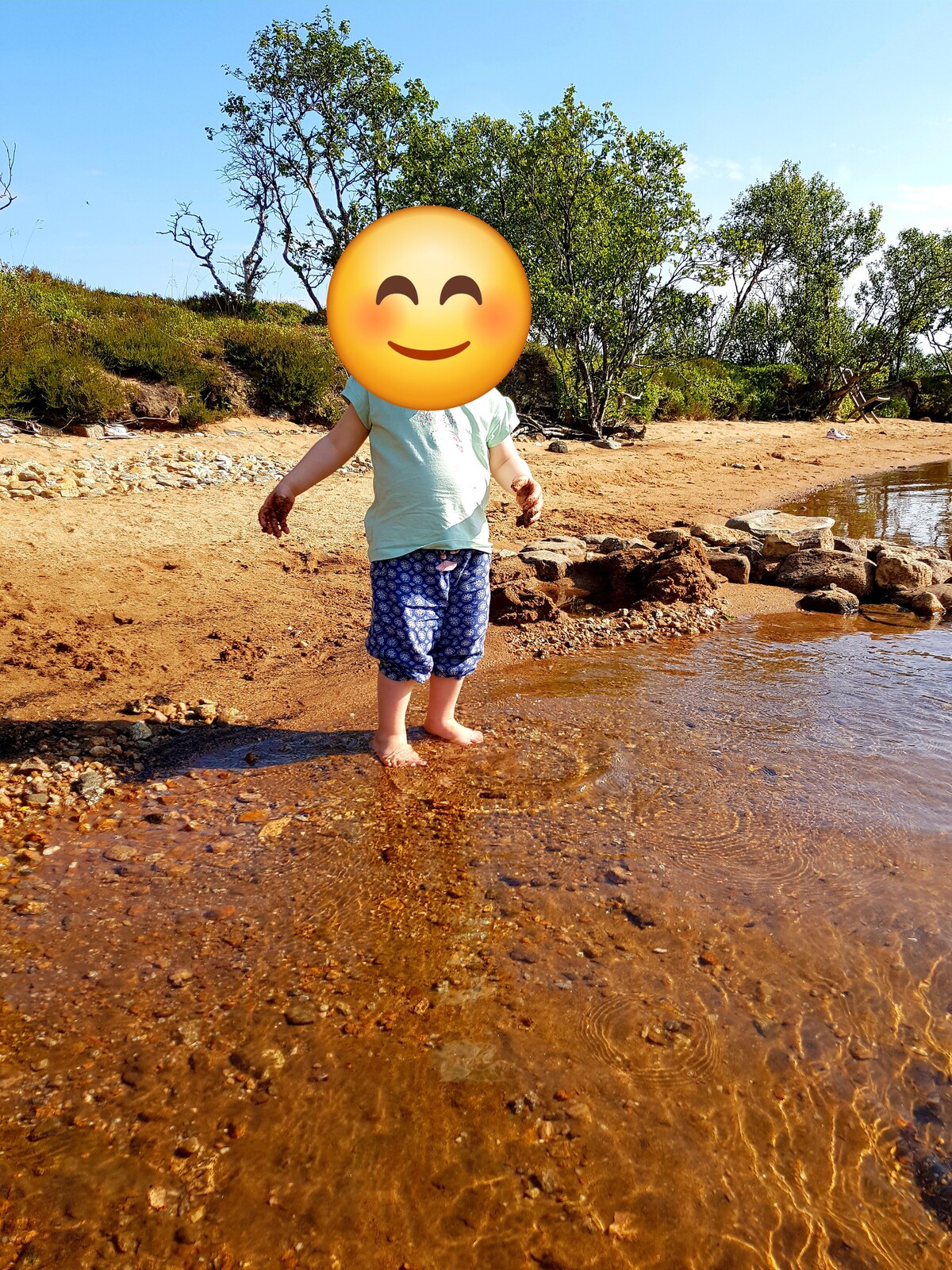 A child stands in shallow water, barefoot on the sandy shore. The surrounding landscape features scattered rocks and low vegetation, with a clear blue sky overhead. Sunlight reflects off the water, creating a warm and inviting setting for outdoor activities.