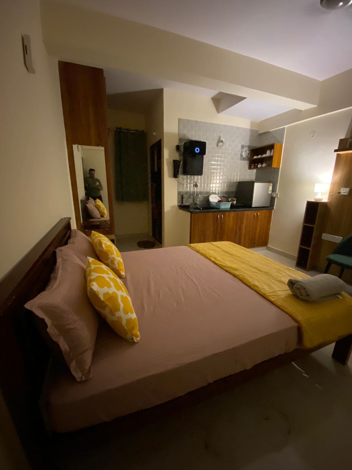 A cozy sleeping area features a spacious bed dressed in soft linens with decorative pillows. A kitchenette with modern appliances is visible on one side, while a full-length mirror reflects the room's layout. Warm lighting enhances the inviting atmosphere.