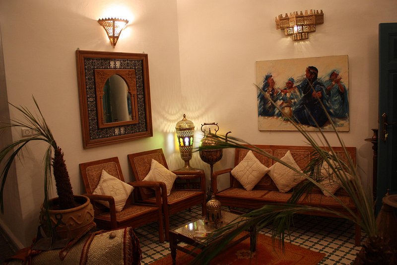 An inviting seating area features traditional Moroccan furniture, including wicker chairs with decorative cushions. A large mirror reflects the warm light from ornate lamps, and a vibrant artwork adds color to the wall. Potted plants enhance the natural feel of the space.