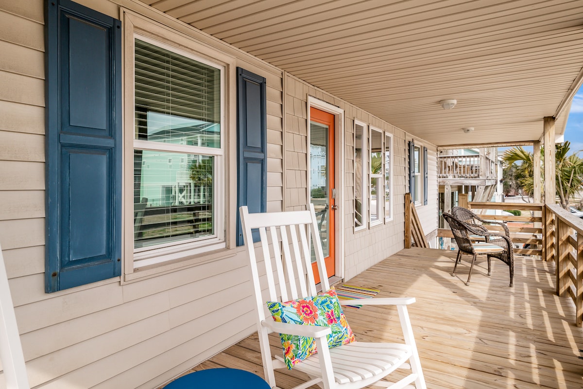 A spacious front porch is presented, featuring a rocking chair and a small blue side table. Colorful cushions add a pop of color to the seating. Tall blue shutters frame the windows, while the entrance door is brightly painted in orange, contributing to a cheerful exterior.
