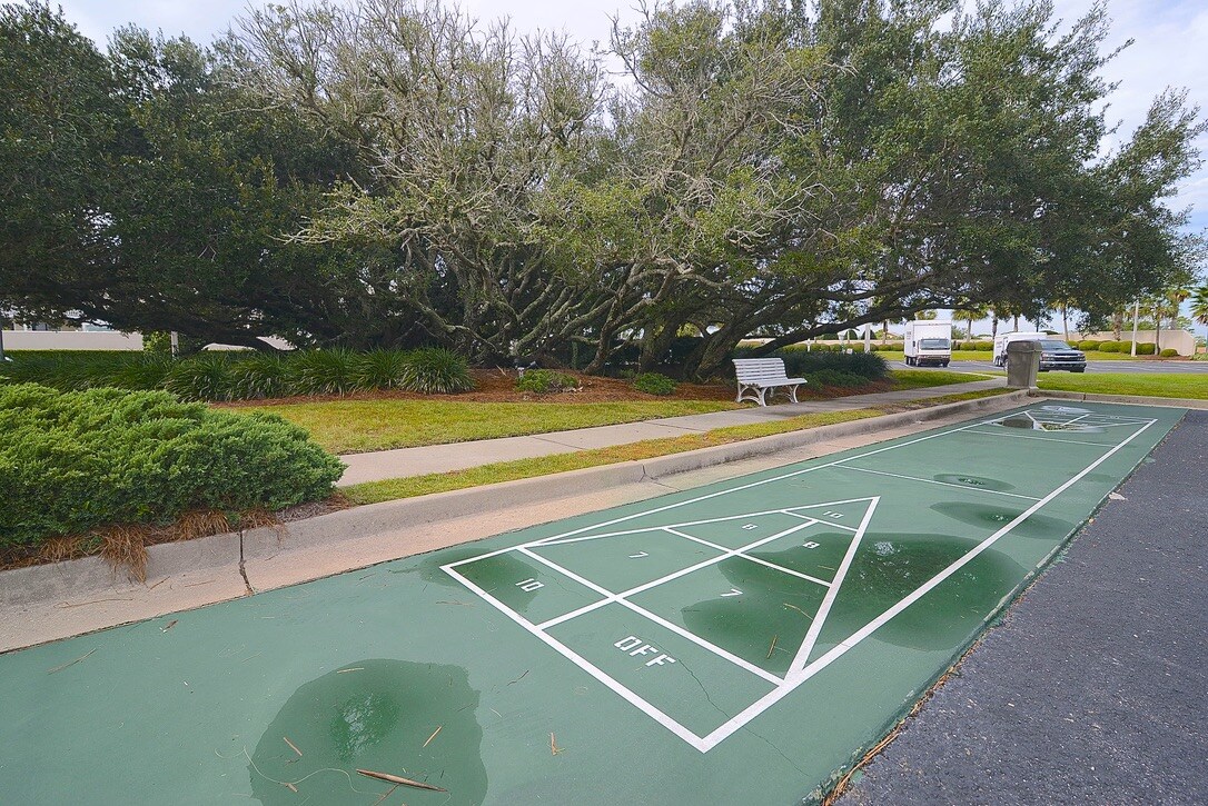 A shuffleboard court is visible on a smooth green surface, marked with game lines. Surrounding the area, trees provide shade, and a nearby bench offers a resting spot. Lush landscaping borders the court, contributing to a serene outdoor environment.