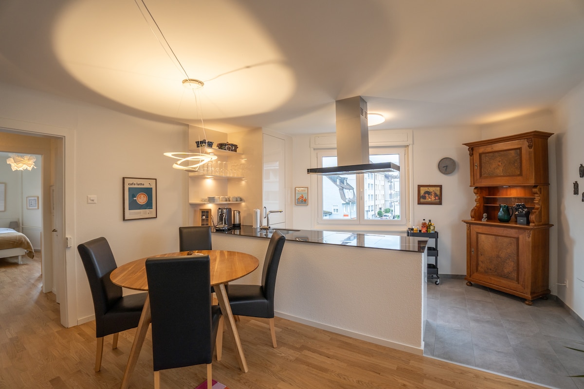 A modern kitchen and dining area are highlighted by a round wooden table surrounded by black chairs. Natural light enters through large windows, enhancing the sleek cabinetry and stainless steel appliances. A vintage cabinet adds a touch of character to the space.