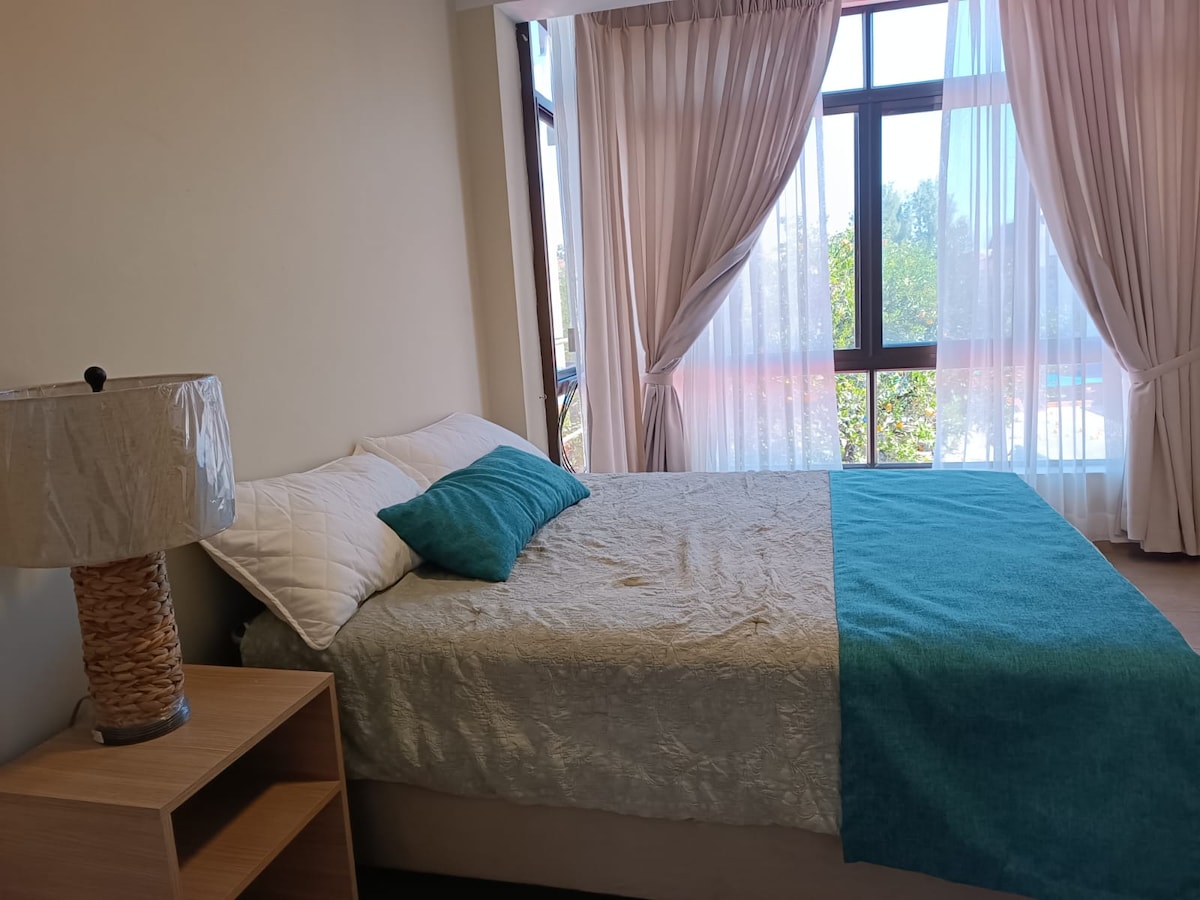 A comfortable sleeping space is presented, featuring a spacious bed adorned with a light-colored quilt and a turquoise throw blanket. Natural light filters through large windows, revealing a view of greenery outside. A bedside lamp adds a subtle touch of warmth.