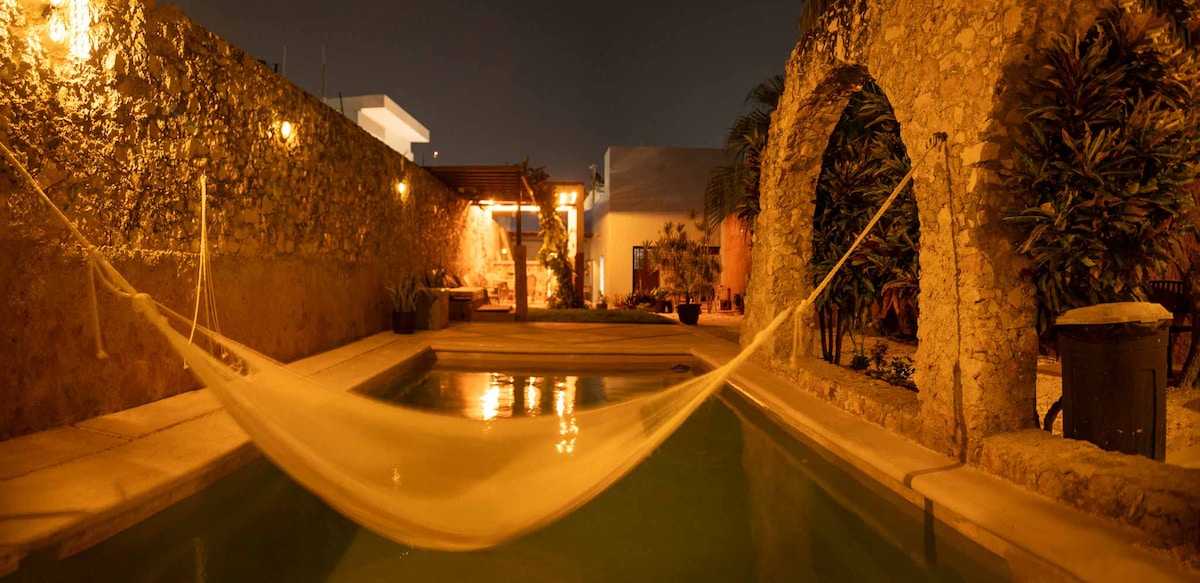 A tranquil swimming pool shimmers under soft evening lights, bordered by lush greenery. A hammock is gently draped between two stone walls, inviting relaxation. The surrounding area features natural stone textures and warm lighting, accentuating the serene ambiance of the outdoor space.