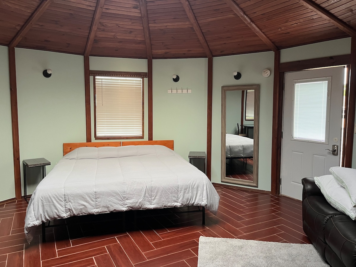 The interior features a spacious sleeping area with a large bed, flanked by nightstands. A full-length mirror reflects the room's design, and natural light enters through the window with blinds. A door leads outside, and a cozy couch is positioned nearby.