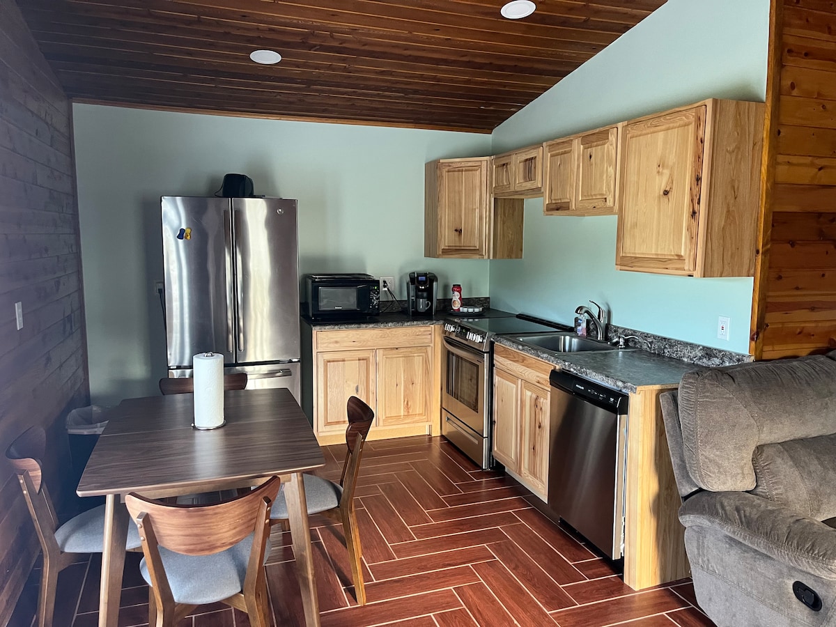 The kitchen area features wooden cabinetry and granite countertops. Modern stainless steel appliances include a refrigerator, microwave, and oven. A dining table with four wooden chairs is positioned nearby, providing space for meals. Soft lighting enhances the open, functional layout.