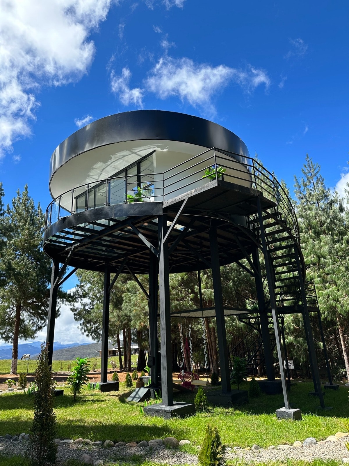 A modern glamping structure, elevated on stilts, features a circular design with large glass windows. A spiral staircase leads to the upper level, surrounded by lush green trees and landscaped gardens, providing a harmonious connection with the natural surroundings.