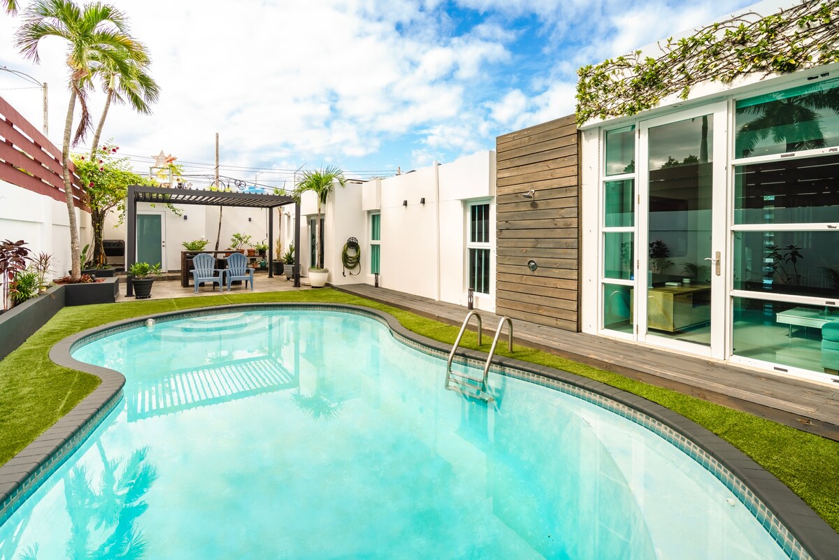 A serene swimming pool stretches across the backyard, framed by lush greenery and wooden decking. Lounge chairs are positioned nearby under a shaded area, providing a relaxing retreat. Large glass doors lead into the home, allowing natural light to illuminate the space.