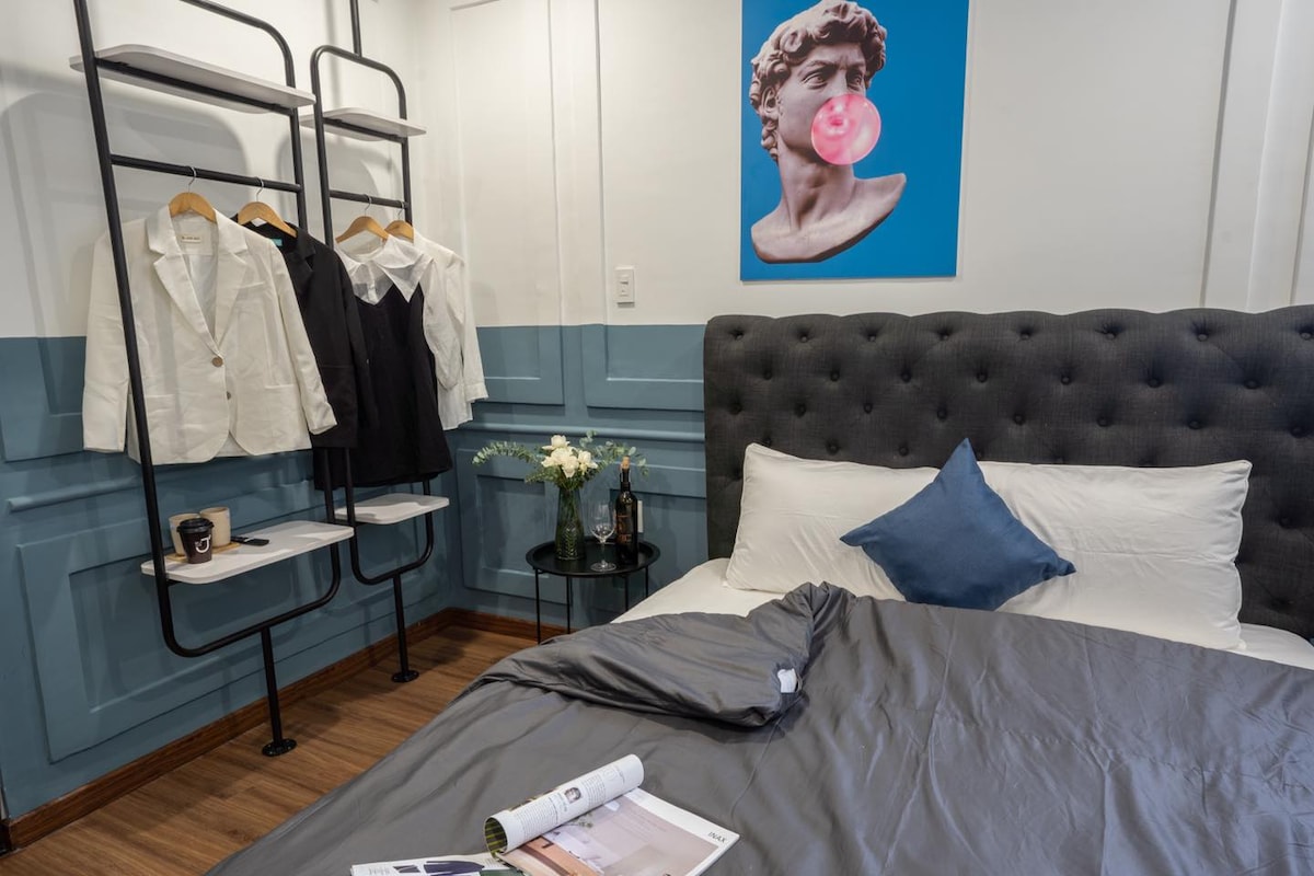 A cozy sleeping area features a tufted headboard and soft bedding in neutral tones. A stylish clothing rack is displayed beside the bed, holding various garments. A small round table is positioned near the bed, adorned with a vase of flowers and casual reading material.