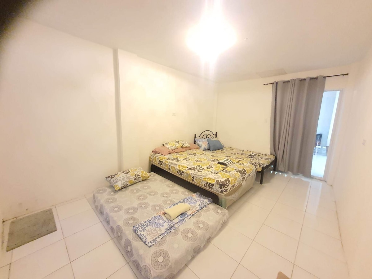 A spacious sleeping area features two beds adorned with patterned bed linens. A smaller mattress is placed on the floor, and soft light is provided by an overhead fixture. Light-colored walls and tiled flooring contribute to the bright ambiance, while a curtain covers a doorway.