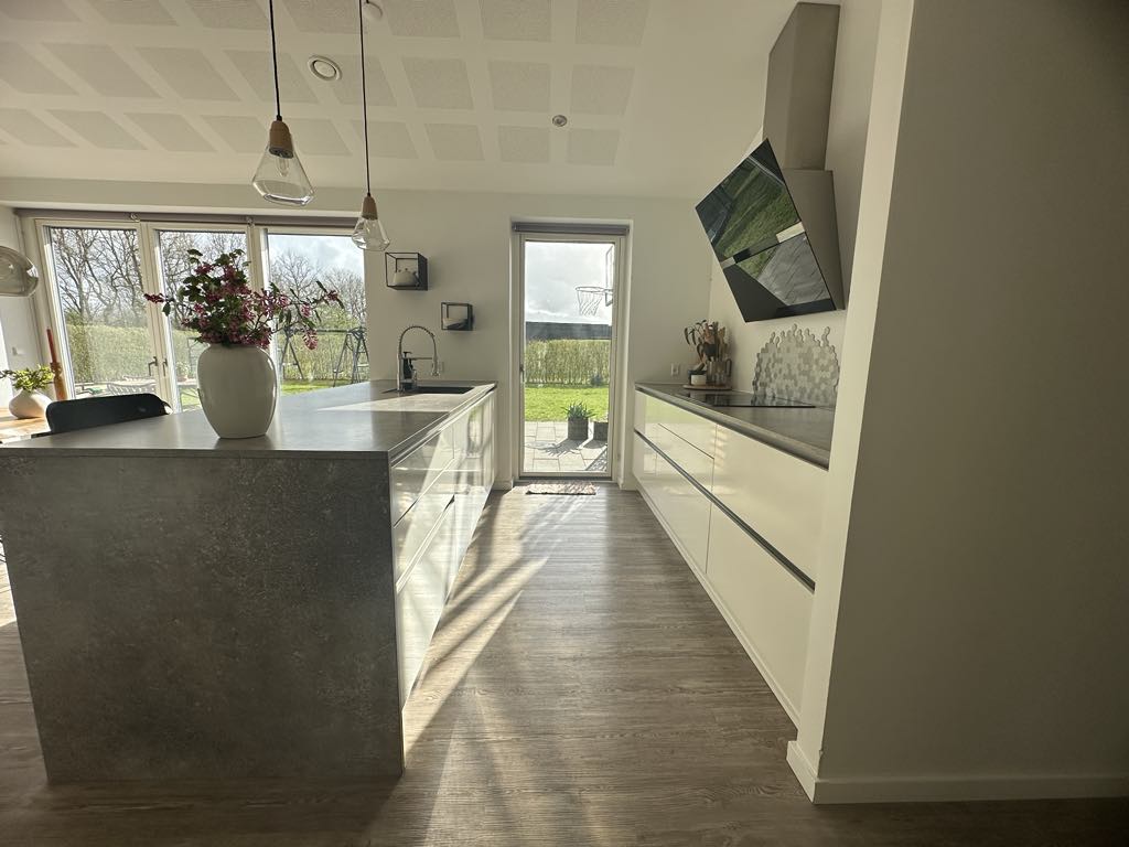 A modern kitchen features sleek white cabinetry and a spacious island with a metallic finish. Natural light floods the area through large windows, with a view of greenery outside. A potted plant and kitchen utensils add a touch of warmth to the contemporary design.