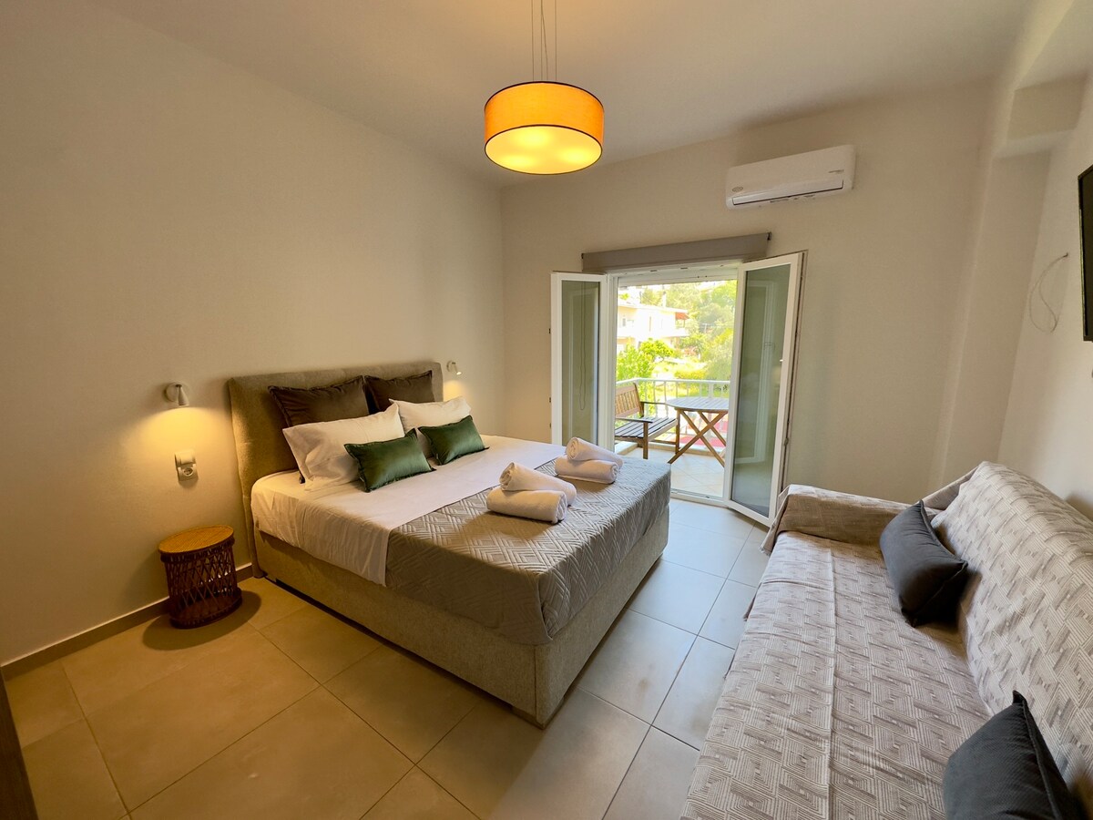 A bright sleeping area is furnished with a spacious bed, adorned with green and white pillows. A comfortable sofa is positioned beside the bed. Light enters through double French doors that lead to a balcony, enhancing the ambiance of the room.