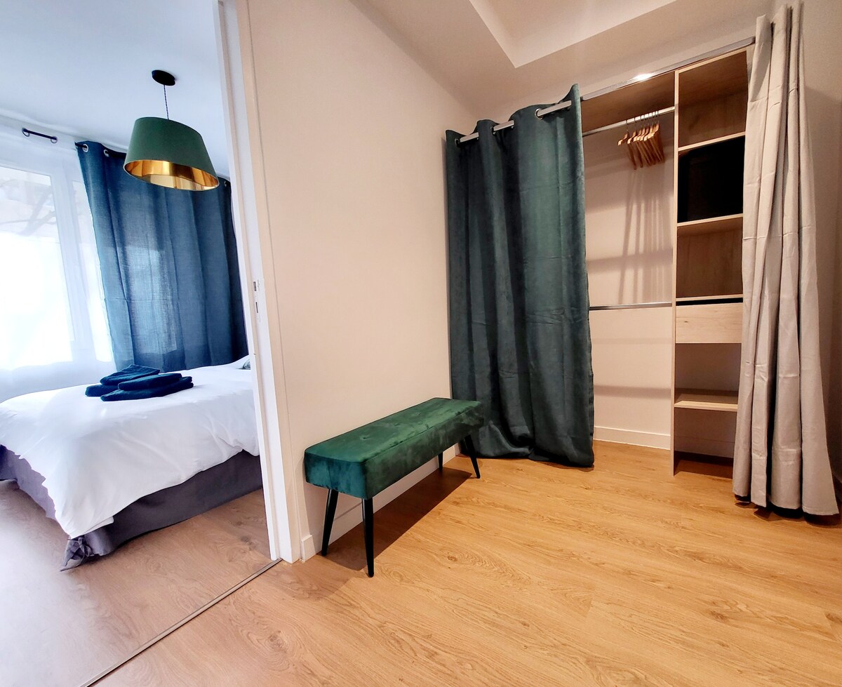 A spacious entryway features a green upholstered bench and a wardrobe with hangers. Light-colored wooden flooring complements the muted tones of the walls and curtains. Natural light filters through the adjacent room, visible through a partially open entrance.