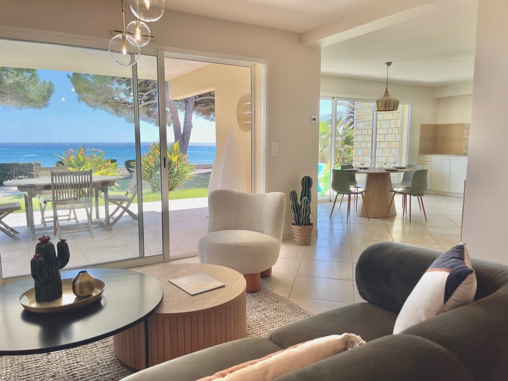 A bright living area features a modern sofa and a round coffee table, with large glass doors opening to a terrace that overlooks the sea. A dining area is visible, showcasing a spacious table and chairs, while greenery can be seen outside.