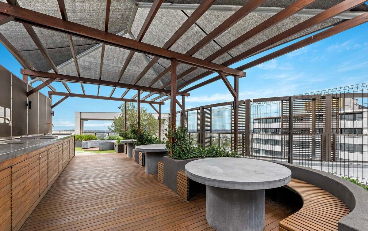 The outdoor terrace features shaded seating areas crafted from natural materials. Circular concrete tables are complemented by bench seating, surrounded by greenery. A spacious wooden deck provides ample space for relaxation and gatherings, with panoramic views of the surrounding area visible in the background.