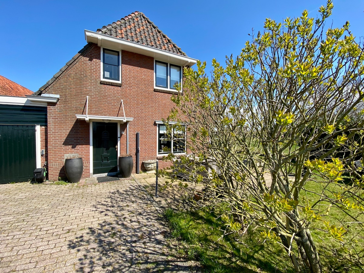 The exterior of a charming two-story villa is depicted, featuring a red brick facade and a gabled roof. Lush greenery surrounds the entrance, with large planters positioned beside the door. The driveway is lined with cobblestones, leading to the quiet surroundings of the property.