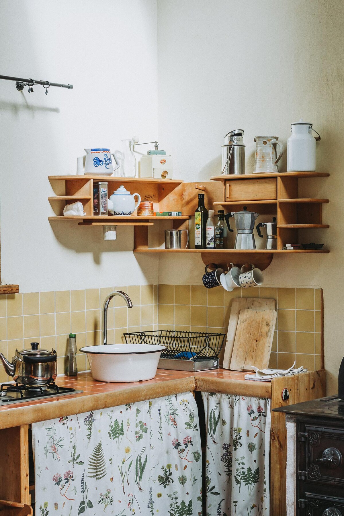 A functional kitchen area is presented, featuring a wooden countertop and a simple sink. Shelves above display an assortment of containers, utensils, and a kettle. A patterned curtain drapes below the counter, complemented by a classic stove visible to the right.