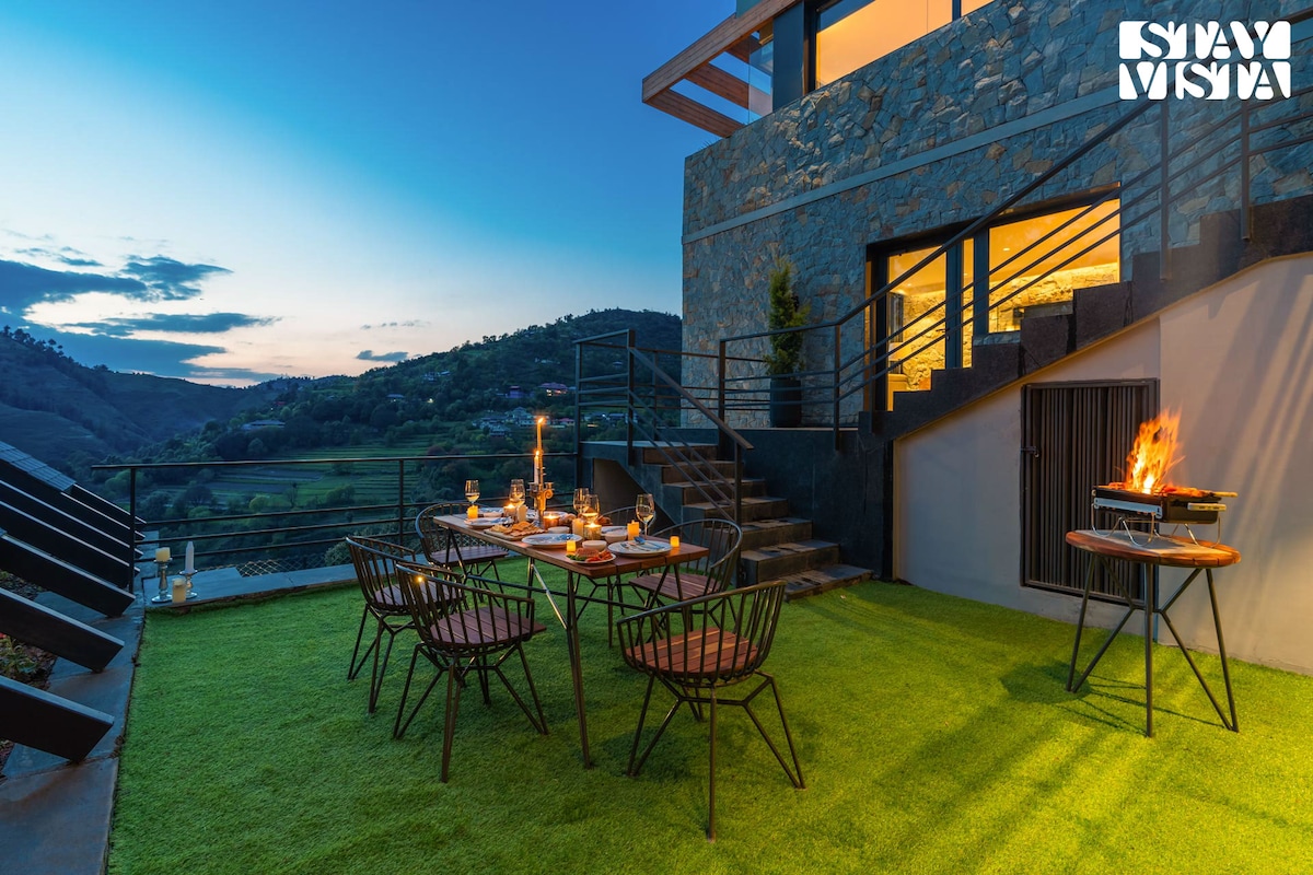 An outdoor dining area features a table set for guests, surrounded by black metal chairs. A barbecue grill sits to the side, with the backdrop of rolling hills and a twilight sky, highlighting the serene ambiance of the estate.