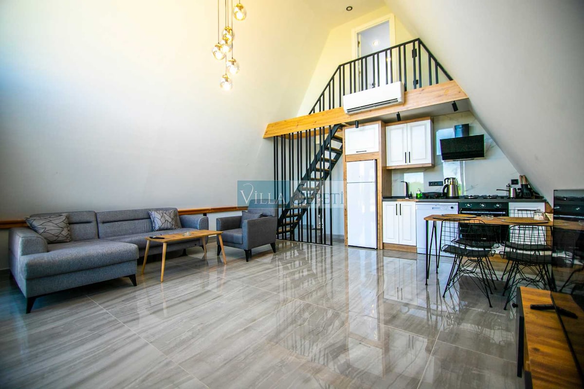 An airy living space features a high ceiling and a mix of modern furnishings. A gray sectional sofa and wooden coffee tables are arranged on a tiled floor. The kitchen area, equipped with appliances, is visible in the background. A staircase leads to a lofted area.