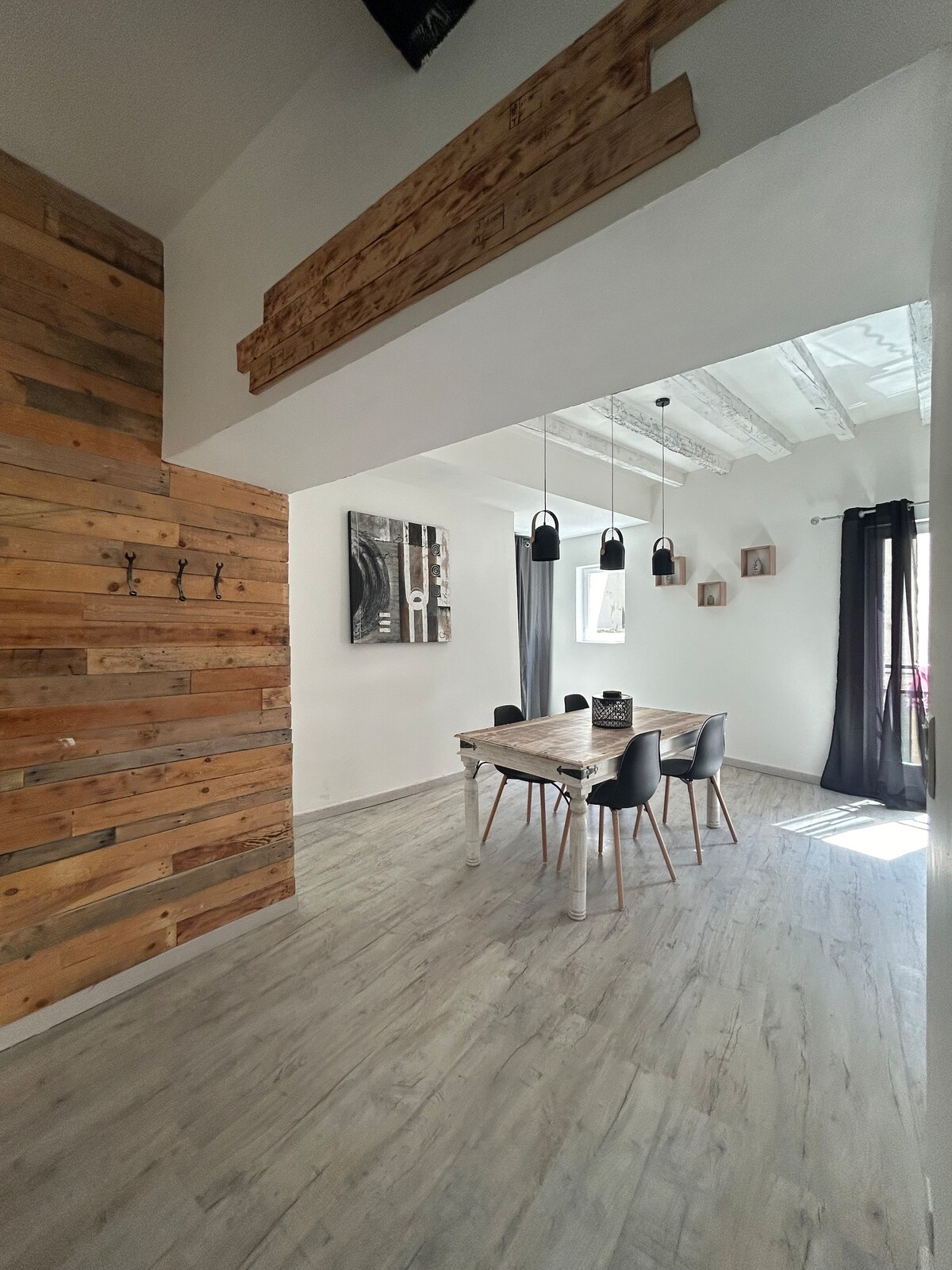 A well-lit dining area is depicted, featuring a wooden table surrounded by several black chairs. The rustic wooden accent wall adds texture. Above, three pendant lights hang from the ceiling, providing illumination. Sunlight enters through a nearby window, highlighting the room's spaciousness.