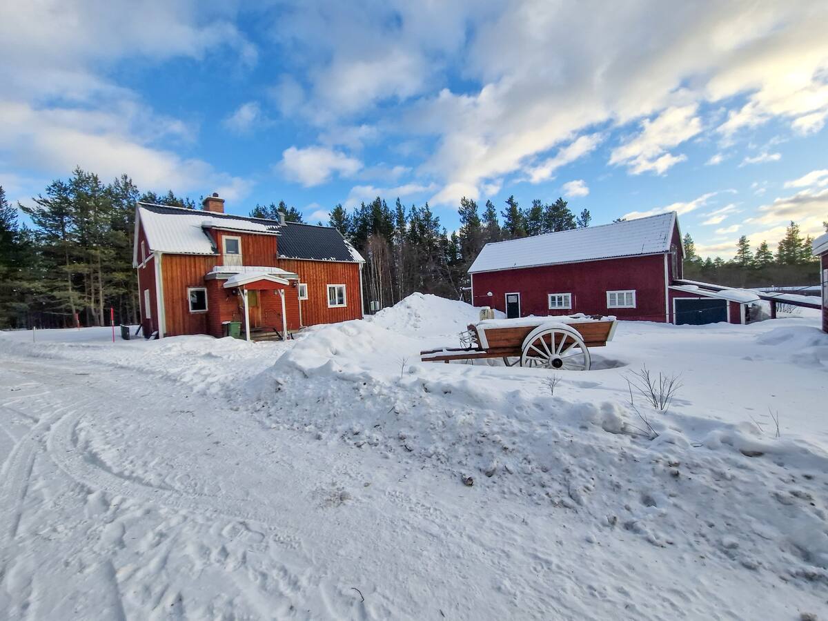 A traditional Swedish red house stands amidst a snowy landscape, surrounded by tall green trees. A wooden cart is positioned in the snow-covered foreground, while a second building is visible to the right, creating a rural, serene setting.