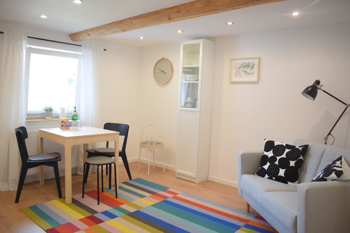 A bright living area features a light wooden beam ceiling and natural light from a window. A modern white table is accompanied by two black chairs and a colorful rug. A gray couch with patterned cushions and a glass display cabinet add to the inviting atmosphere.