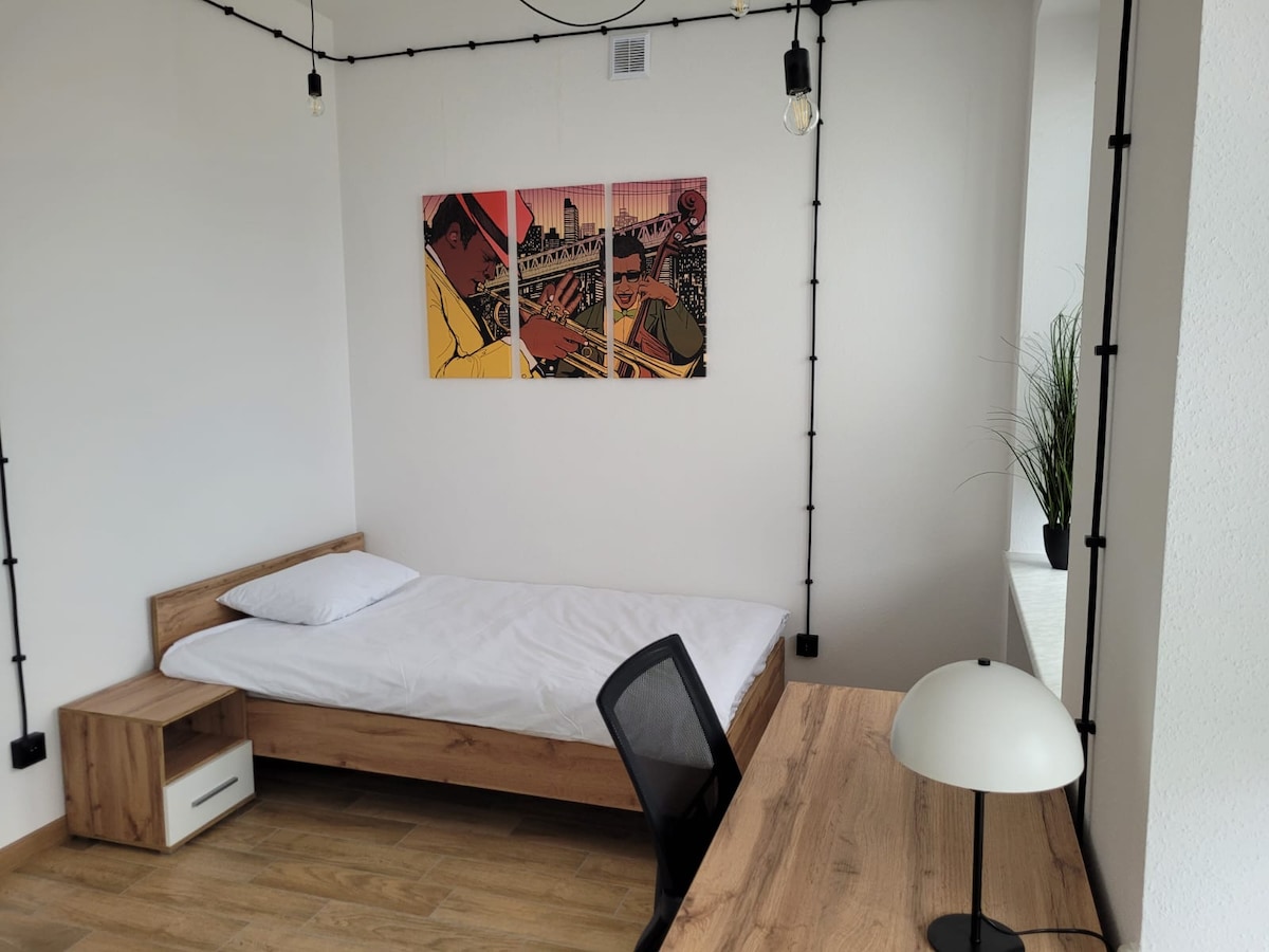 The room features a minimalist design with a wooden bed and a simple mattress. A small nightstand and a workspace with a desk chair are included. A wall art piece adds color, while a potted plant sits on the windowsill, enhancing the bright, airy feel of the space.