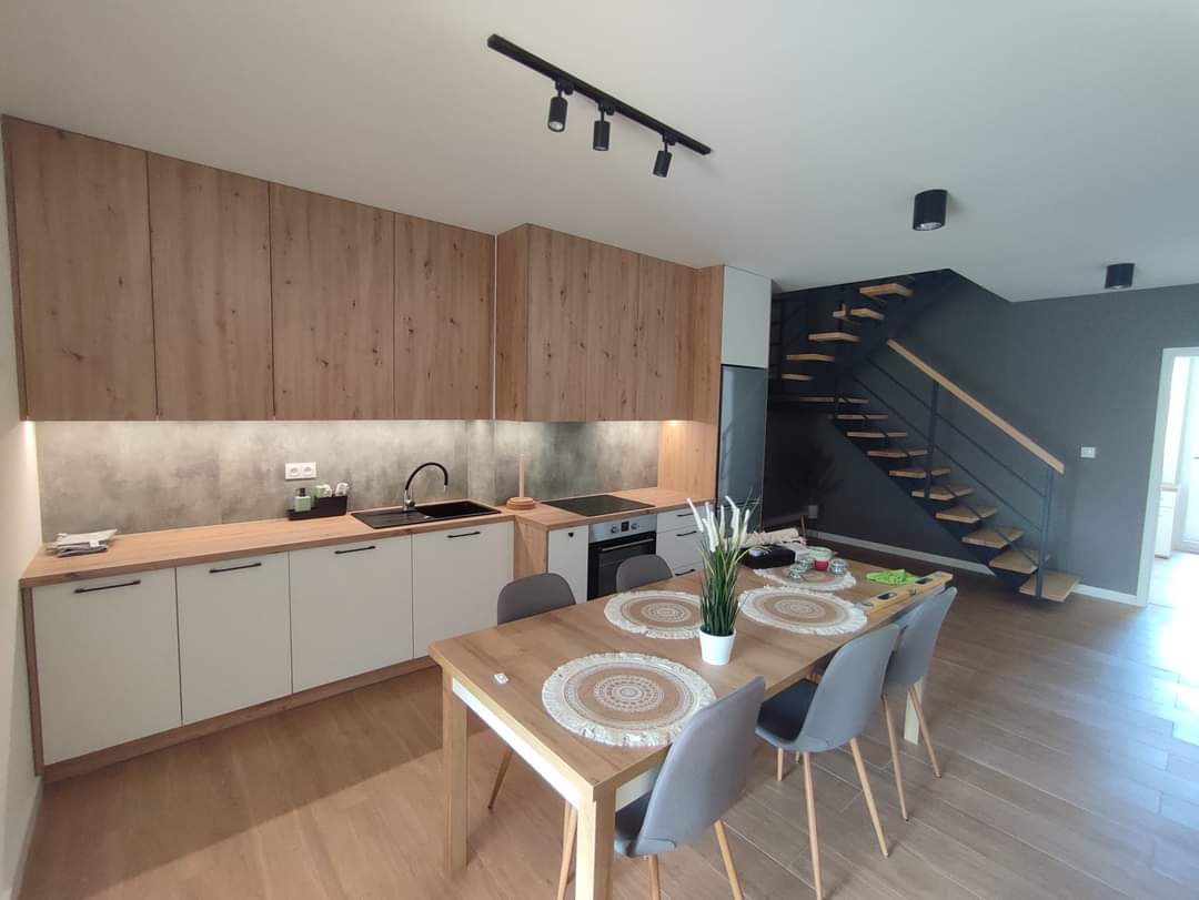 A modern kitchen and dining area features wooden cabinetry and a sleek countertop. The circular dining table is surrounded by grey chairs, and houseplants add a touch of greenery. A staircase with wooden steps and black railings is visible in the background.