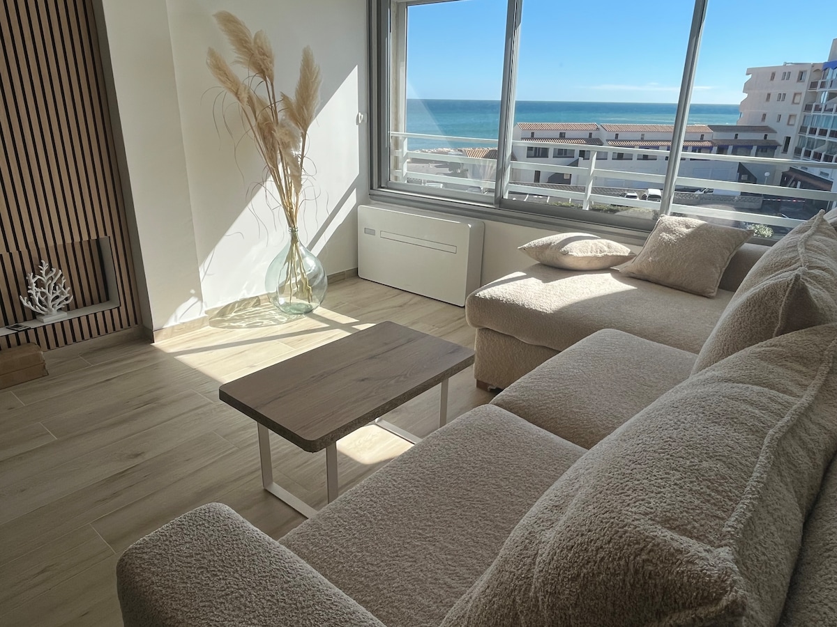 A sunlit living area features a light-colored sectional sofa positioned beside a small wooden coffee table. A large window offers a view of the sea, while a decorative plant in a glass vase adds a natural touch to the space.
