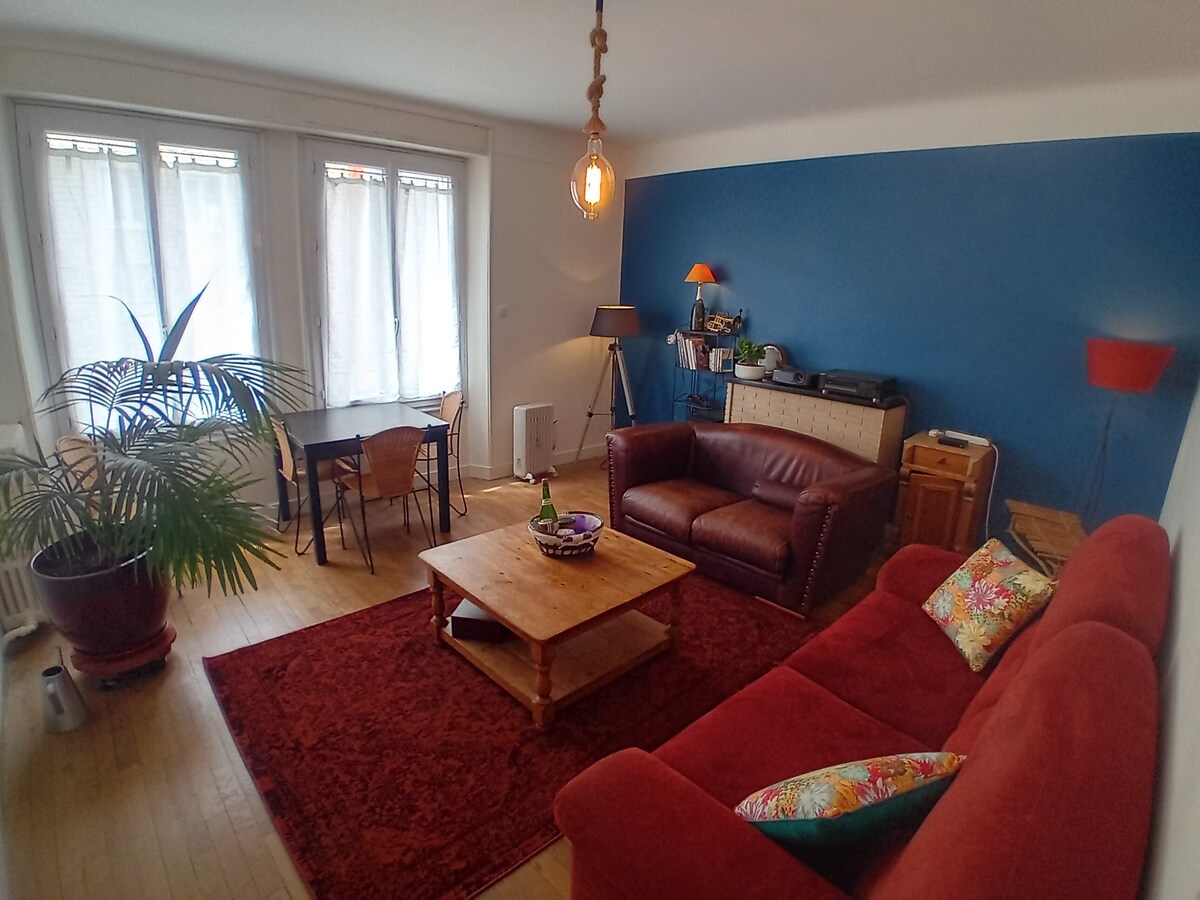 A spacious living area features two comfortable sofas and a coffee table placed on a vibrant red rug. A dining table with four chairs is visible, along with a tall plant. Natural light enters through large windows covered with sheer curtains.