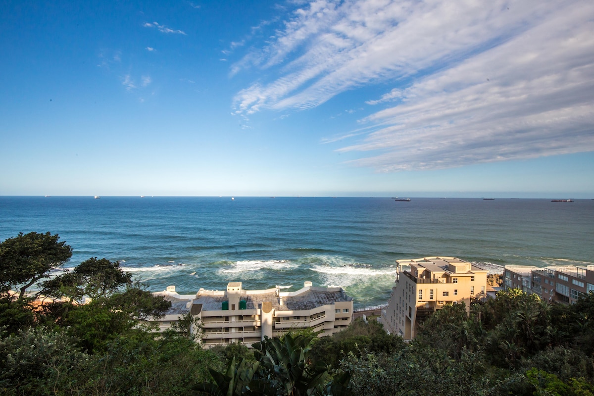 A sweeping view of the ocean stretches across the horizon, showcasing varying shades of blue and white from waves. The coastline features a mix of residential buildings, interspersed with greenery, under a sky adorned with gentle clouds.