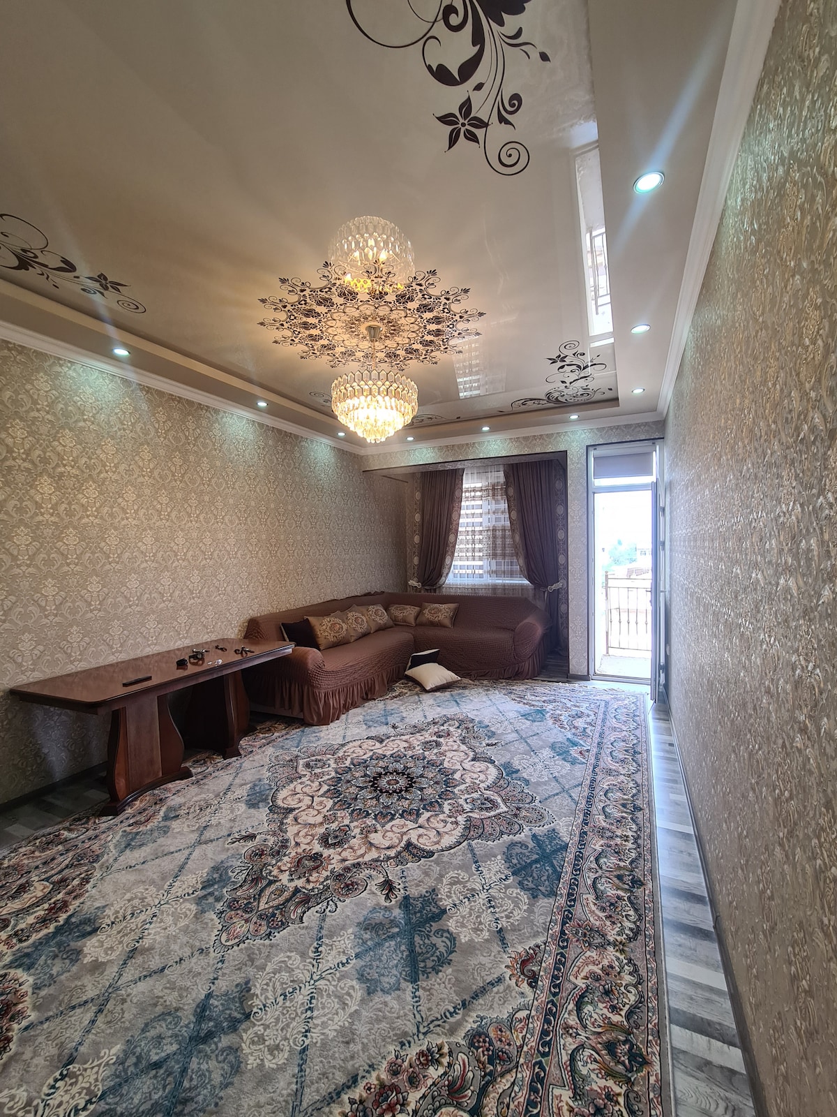A well-lit living area features ornate ceiling designs and a sparkling chandelier. A plush sofa is positioned alongside a wooden table. The space is enhanced by a large, intricately patterned area rug, with a glass door leading to a balcony, allowing natural light to enter.