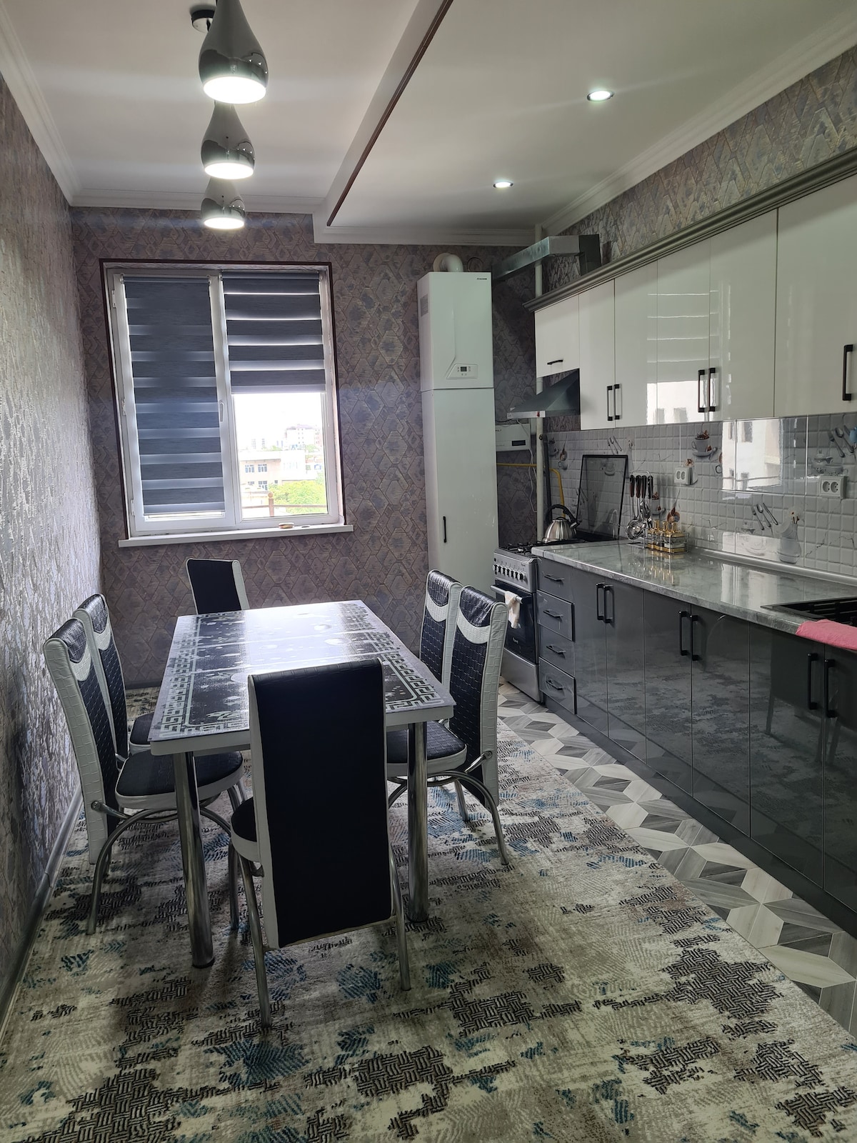 A modern kitchen is showcased with a sleek, glossy cabinet design and a long dining table surrounded by six chairs. A patterned rug covers the floor, while large windows allow natural light to brighten the space. Stainless steel appliances and a minimalist backsplash complete the contemporary look.