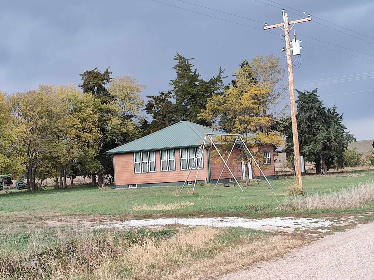A historic schoolhouse is nestled among trees, featuring a green metal roof and large windows. An empty swing set stands in the grassy area next to the building, with tall trees providing a natural backdrop. A gravel road leads to the property.