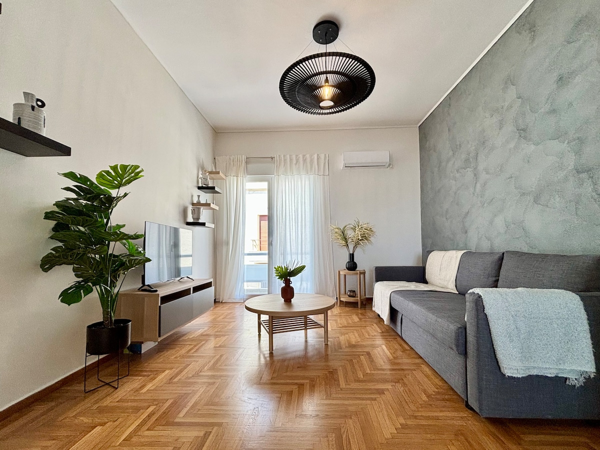 A living area is presented with a modern sectional sofa positioned beside a coffee table. A flat-screen television is mounted on the wall opposite, while large glass doors allow natural light to enter. Greenery is provided by a potted plant, enhancing the space.