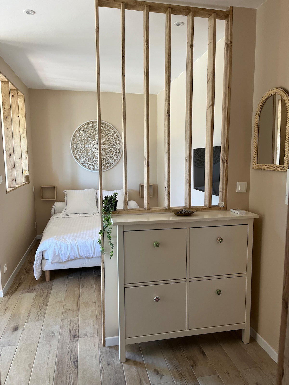 A light and airy bedroom space is visible, featuring a simple bed against a warm-toned wall. A wooden divider adds a touch of elegance, separating the sleeping area from the entryway. A neutral-colored dresser with four drawers sits in the foreground, enhancing the room's functional layout.