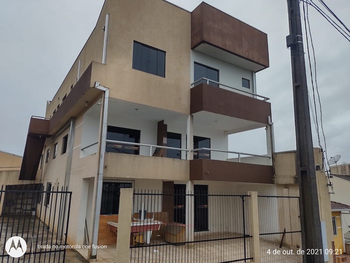 A multi-story building features a modern facade with a combination of neutral and dark-colored panels. Large windows allow natural light to enter, and a balcony is visible on the upper level. The ground level is partially obscured by a fence and other structures.