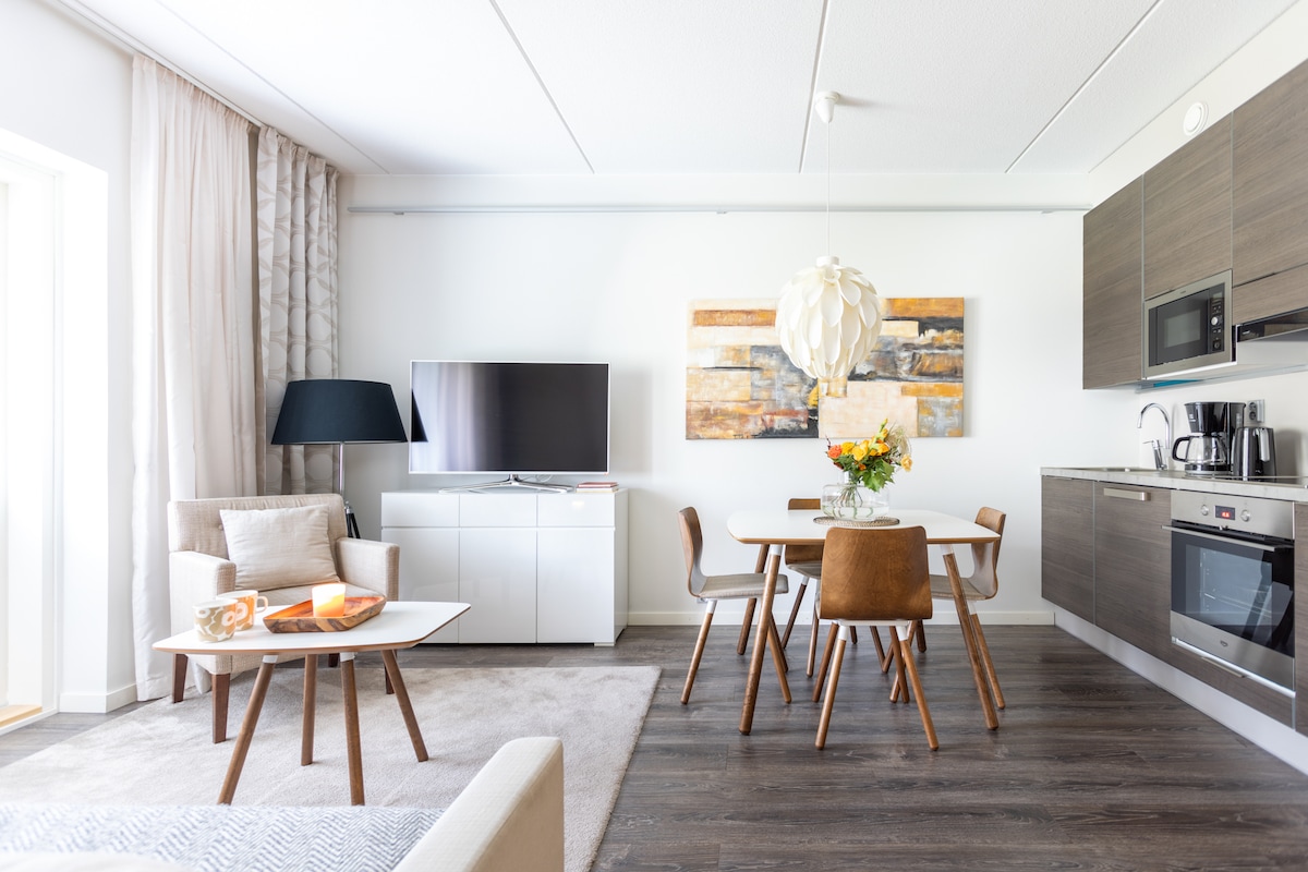 A bright living area is featured, showcasing a comfortable sofa and a small coffee table. A dining table with four chairs is positioned nearby, with a vase of flowers as a centerpiece. A wall-mounted television and modern kitchen appliances are visible, enhancing the functional design.
