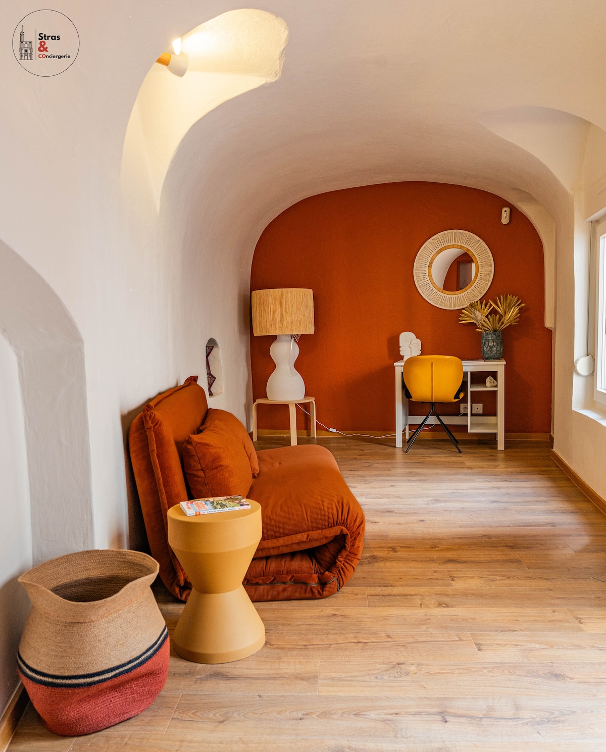 A versatile room features an earthy orange accent wall and a cozy futon. A small desk with a round mirror is positioned against one wall, while a decorative lamp and a simple side table add to the inviting decor. A woven basket sits on the floor.